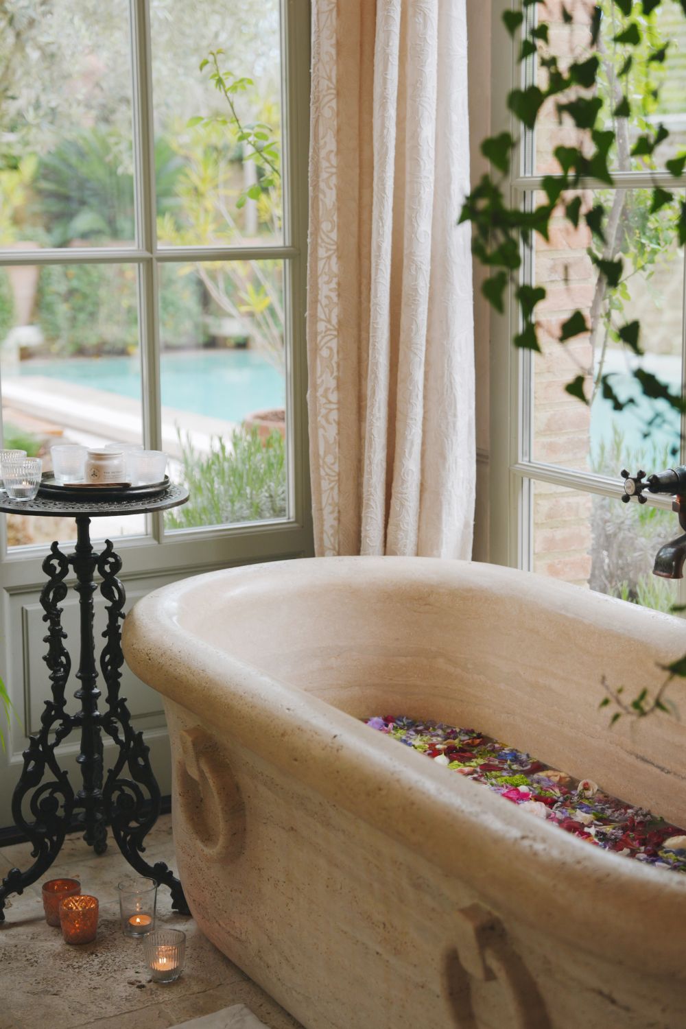 A light stone bath next to a corner sage green window, facing a pool.