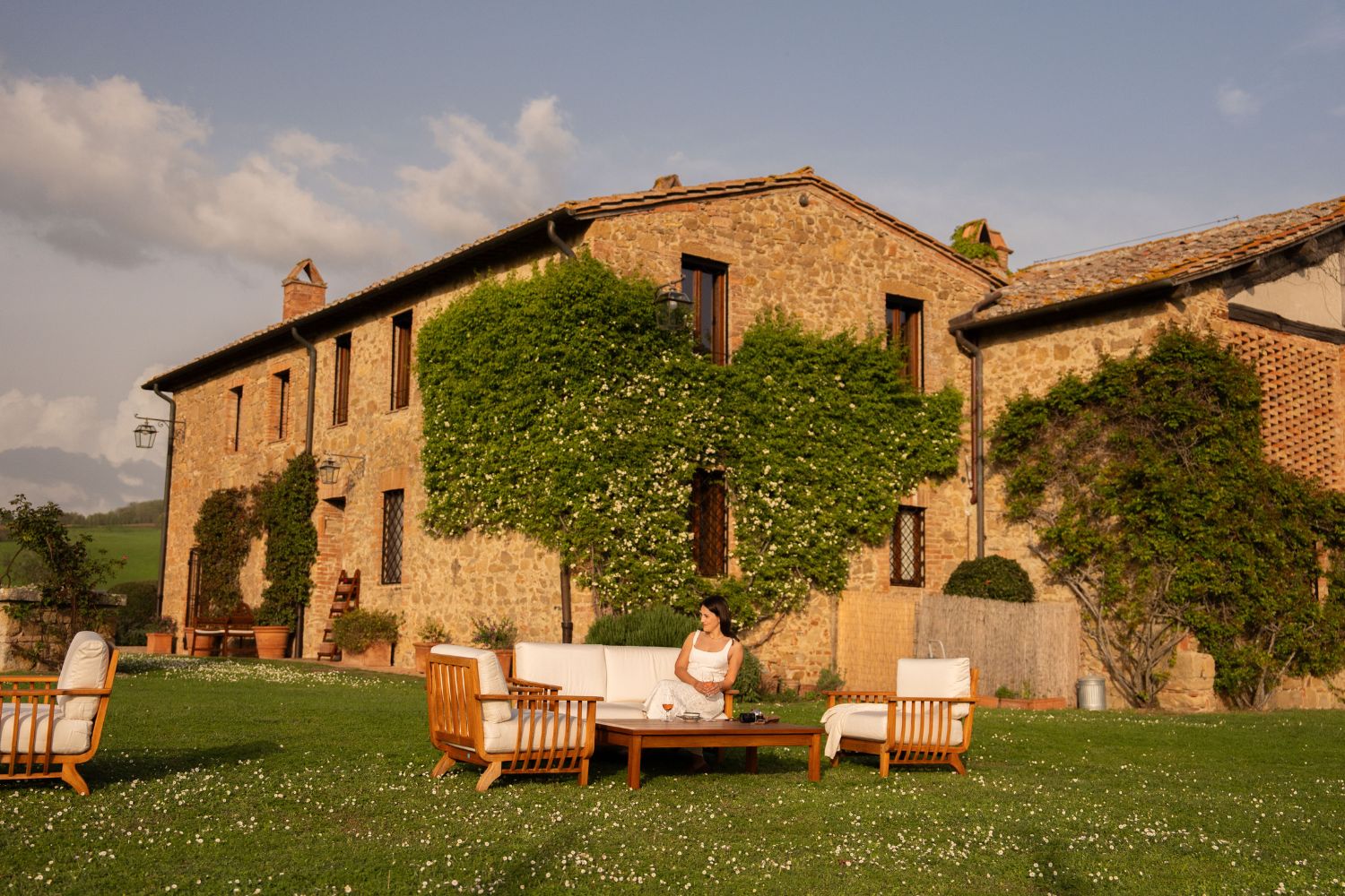 A stone Tuscan house, with climibing flowers and dark wooden windows, alongside a white and wooden outdoor seating where a young lady with a white dress is seating.