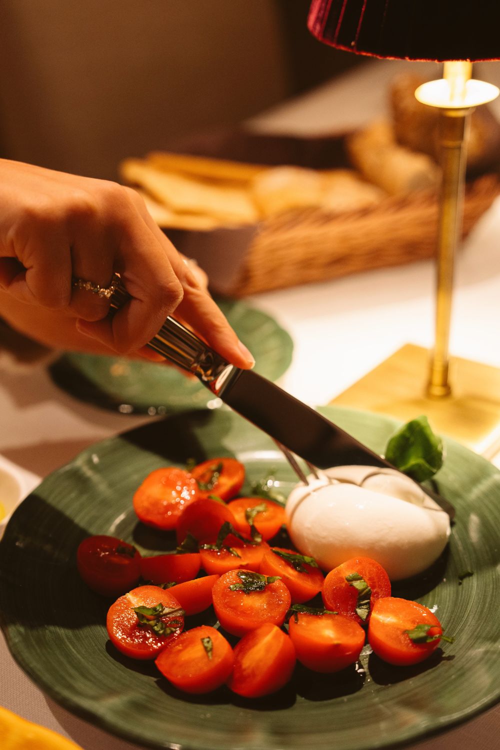 A dark green dish, where a hand is cutting a cheese next to red tomatoes.
