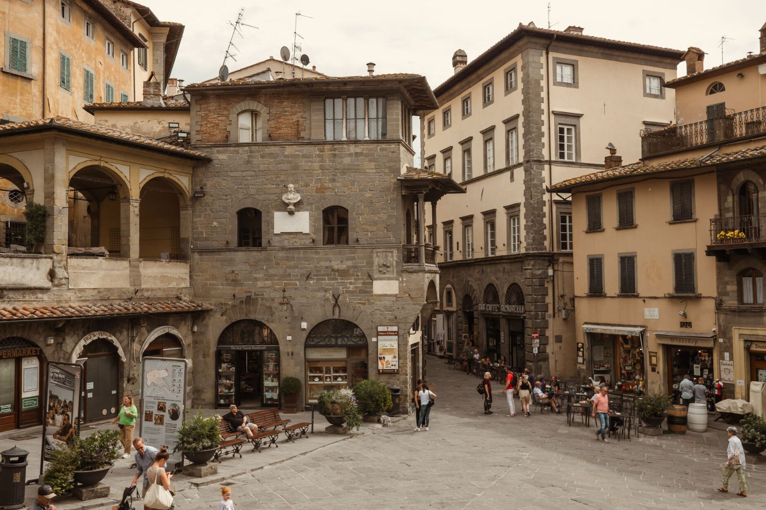 Village centre, with several cobbled stone houses with arches and shutters. Stone floor with benches, tables, vases with flowers and people walking around.