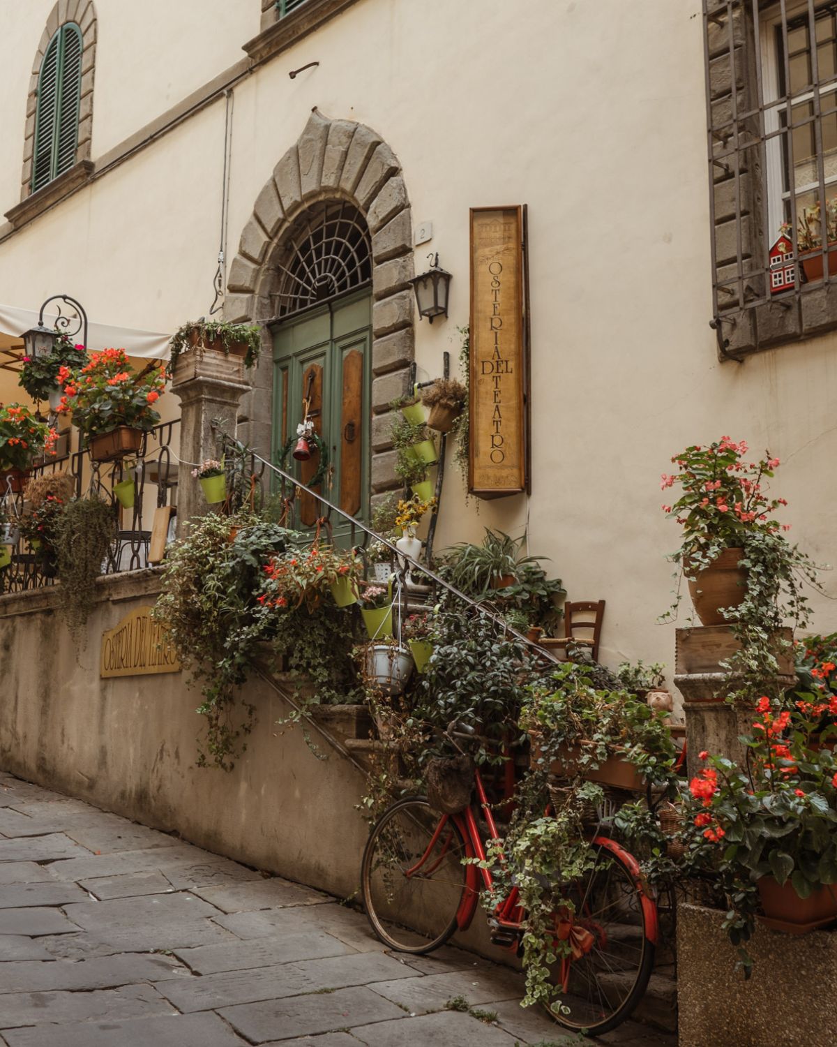 Staircase with hanging vases and plants, next to a red bike. A green door at the top of the stairs with a stone arch on the top.