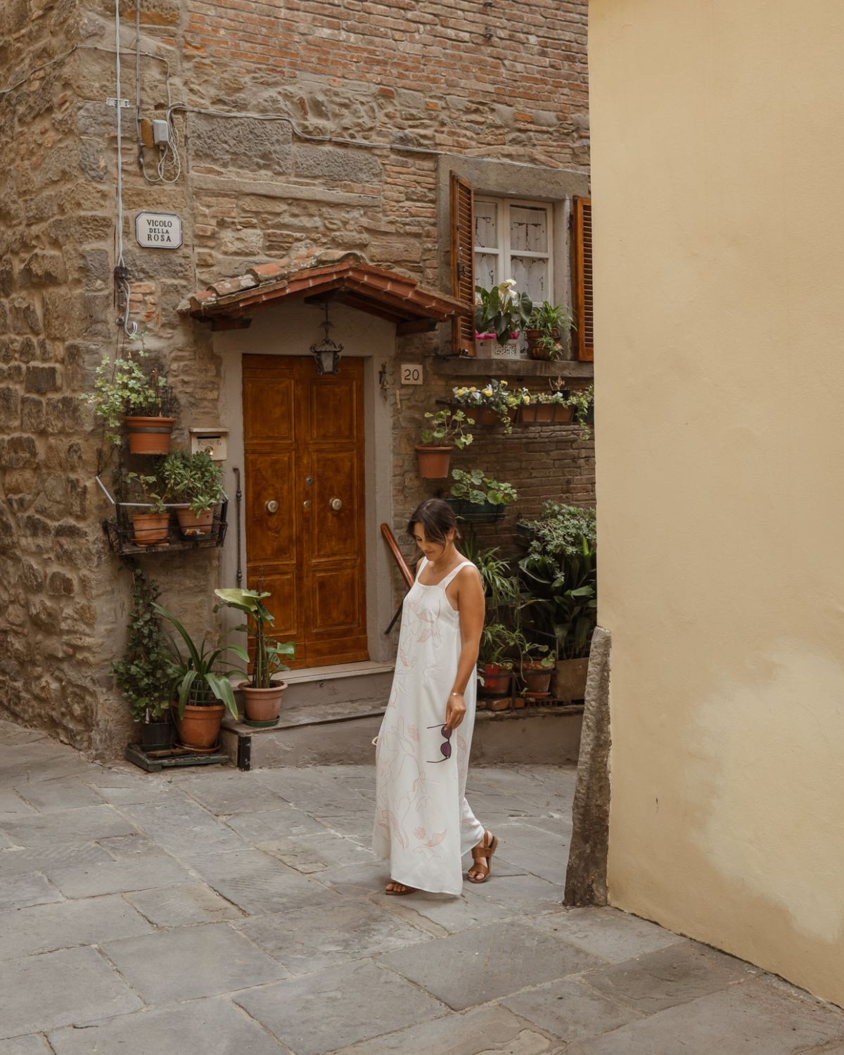 Woman in a white dress, standing in a street with a cobbled stone house, with wooden shutters and a door, next to hanging plants in vases.