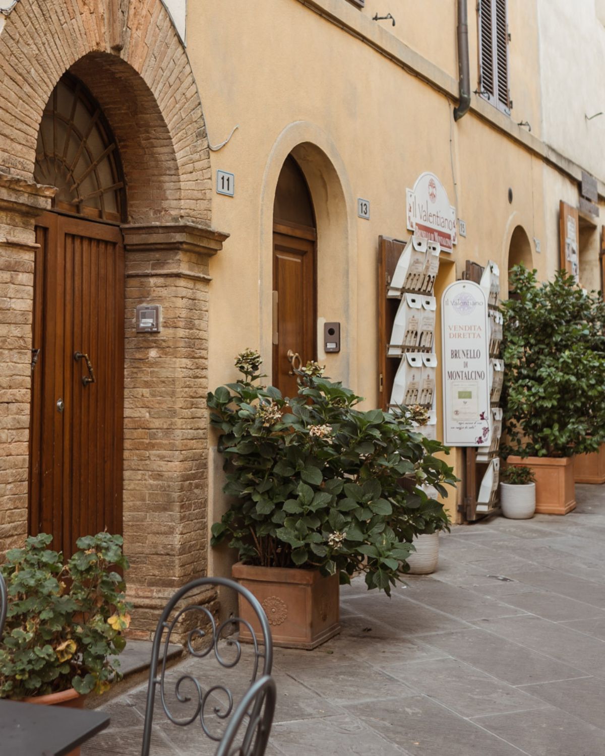 Lively streets with a light orange house, with brown doors, hanging products and vases with plants on the floor, next to a set of iron chairs.
