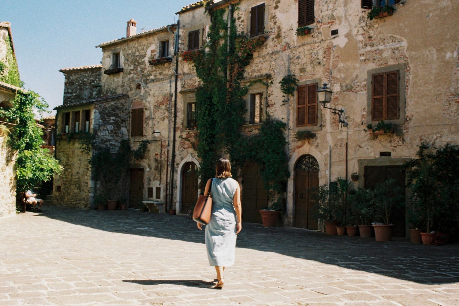 Woman in a light blue dress, walking in a street with cobbled stone houses, with wooden shutters, next to hanging plants in vases.
