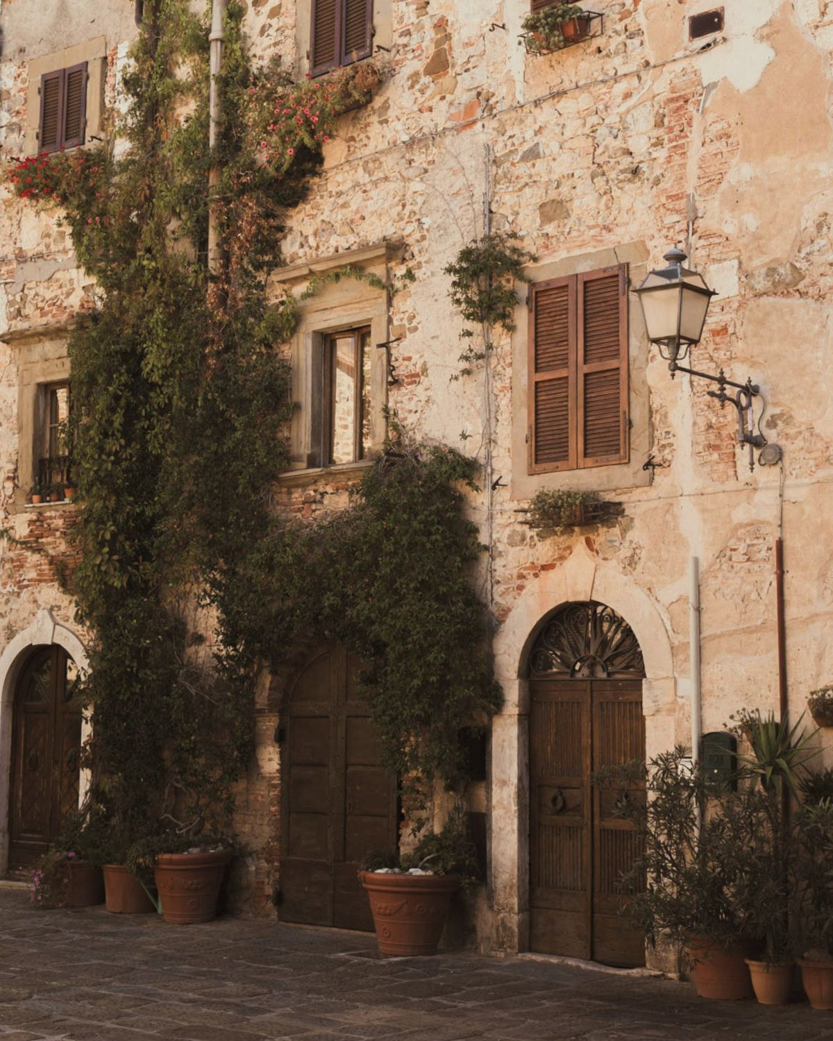 Cobbled stone house, with shuttered windows and several vases with plants on the floor.