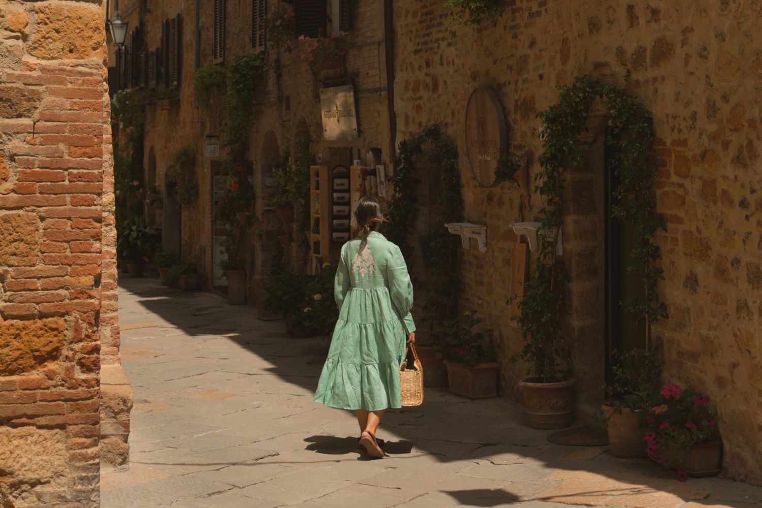 Woman in a teal dress, standing in a street with cobbled stone houses, with wooden shutters, next to hanging plants in vases.