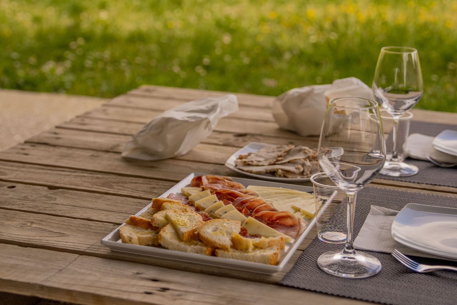 Center wooden table with cutlery, glasses, cheese, wine, water, bread and cured meats.