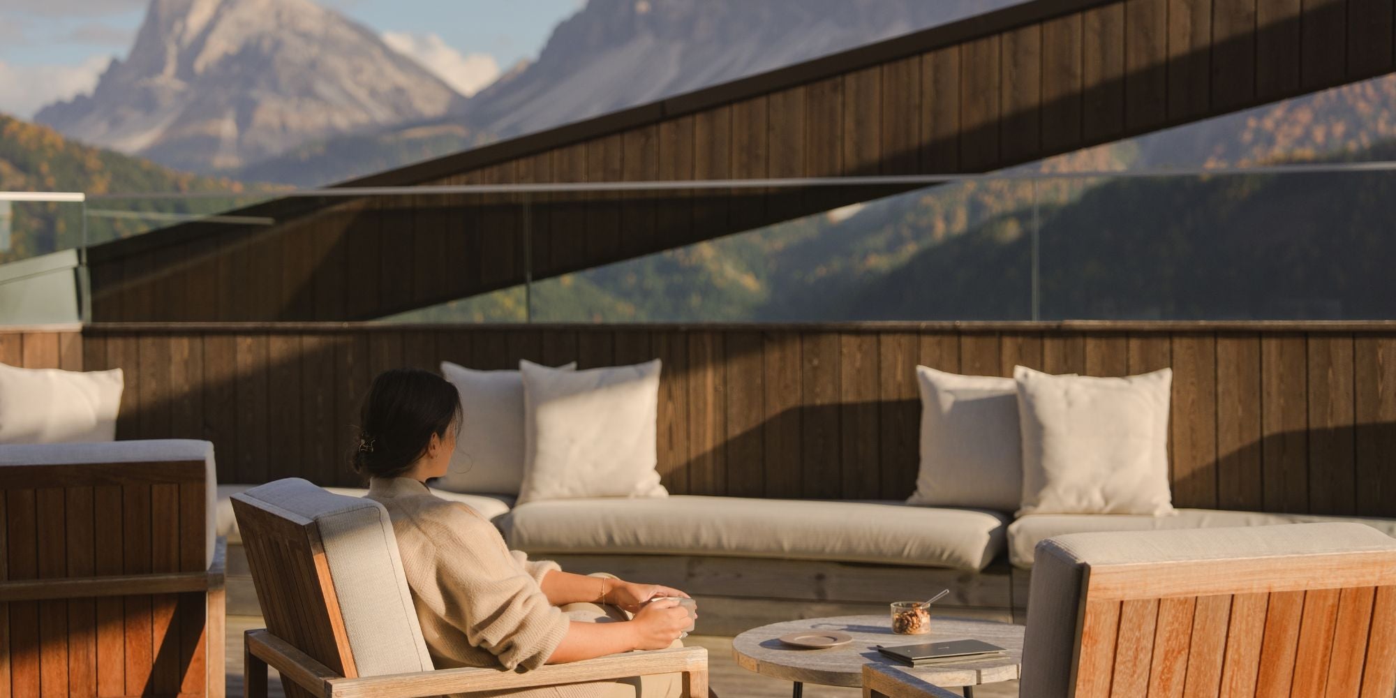 A lady sitting on an outdoor cream chair, on a hotel wooden terrace, overlooking a high mountain.