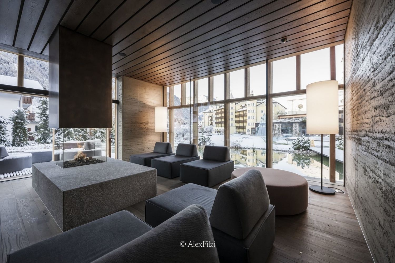 A wood, stone and concrete hotel lounge space, with lounge chairs and a fireplace, surrounded by large windows, overlooking a snowy exterior.