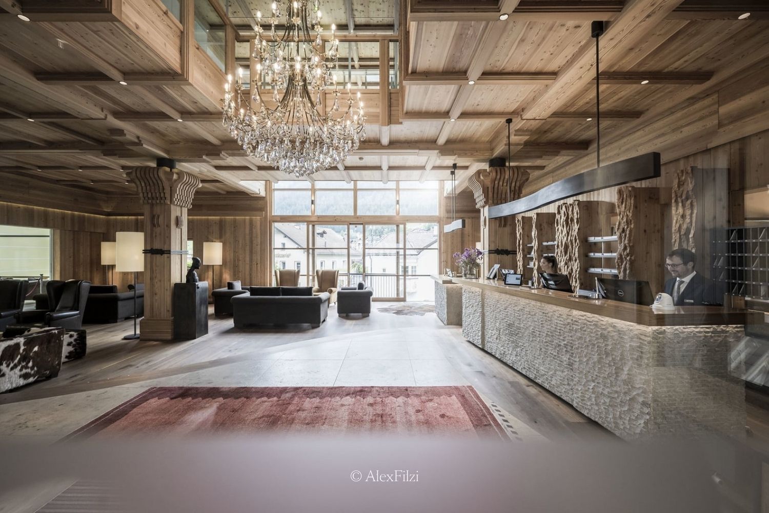A wooden hotel entrance, with a crystal chandelier, a red rug and black furniture, a stone counter where the employees sit behind.