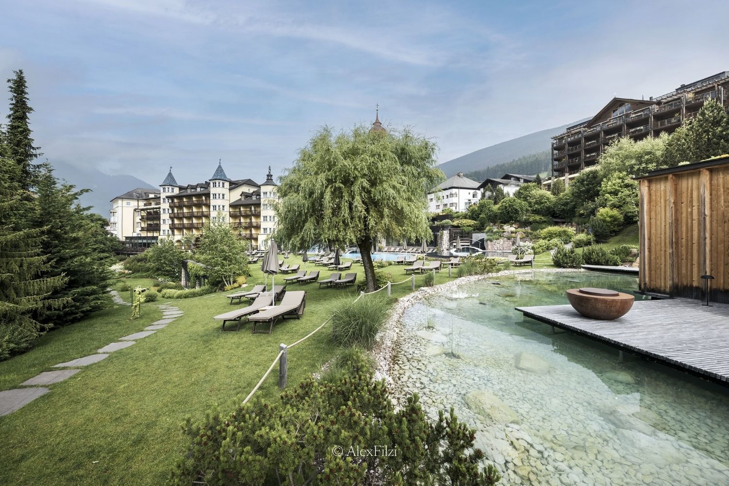 A green outdoor hotel scenery, with a small pond, outdoor sunbeds, three largw buildings and mountains in the back.