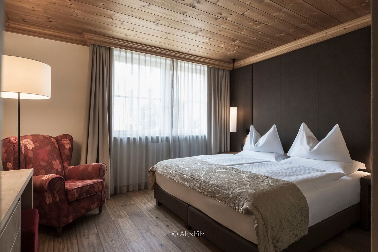 A hotel room, with a dark brown wall, wooden ceilings and floors, a red armchair and white linens.