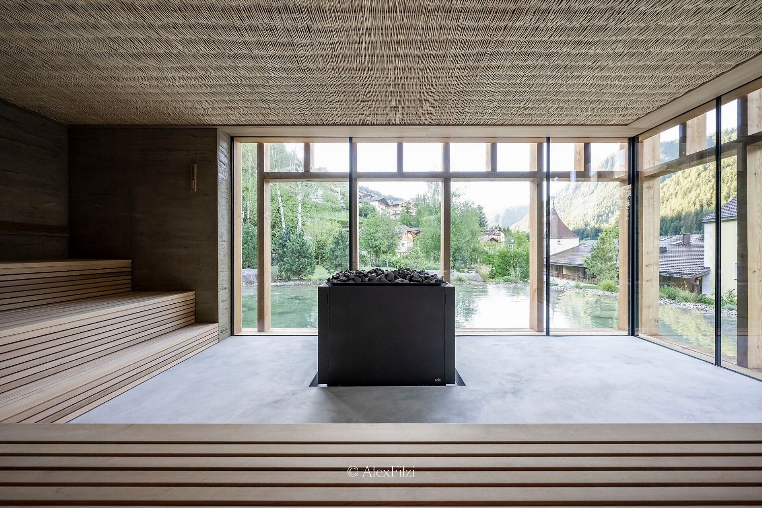 A sauna setting, covered in wood and large windows facing a small outdoor pond, buildings and greenery.