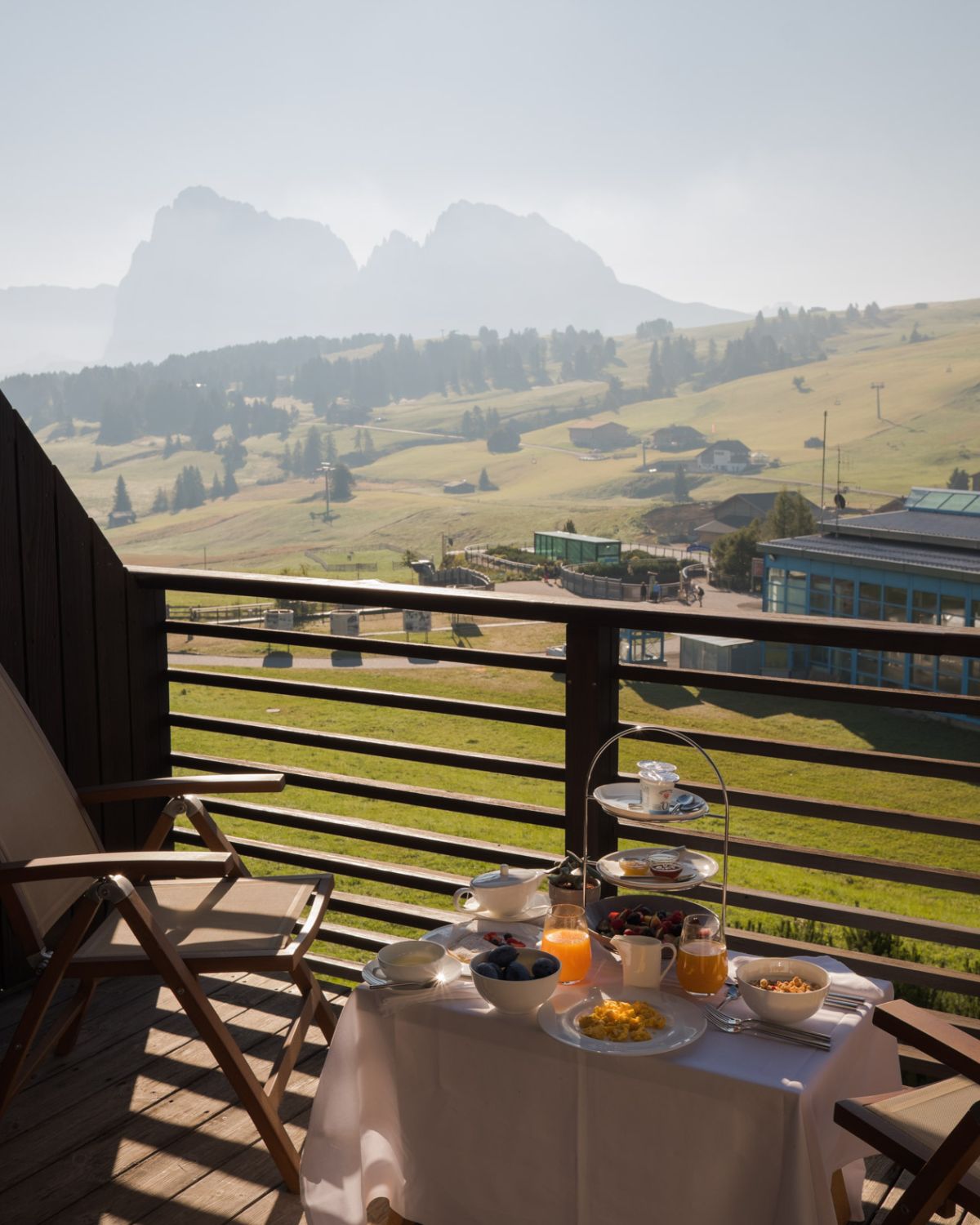 A hotel balcony, with outdoor furniture, breakfast food on the center table, overlooking a green hill and mountains.