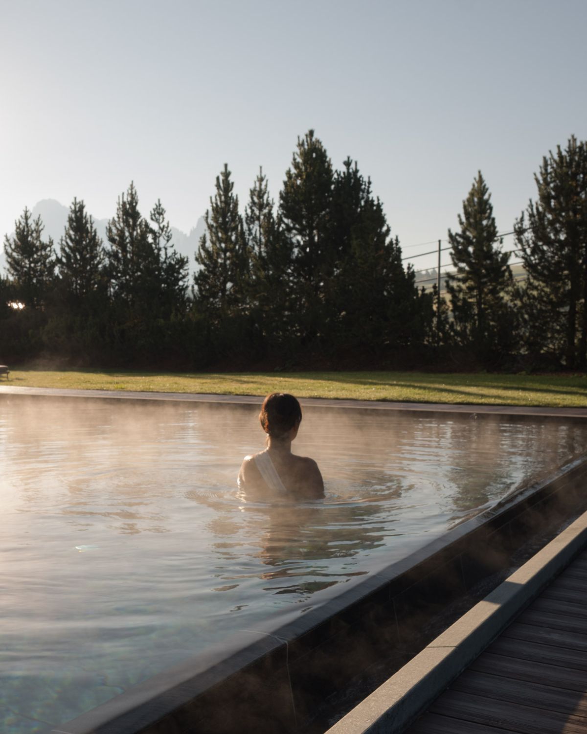 A lady inside an outdoor heated pool, overlooking high trees and mountains.