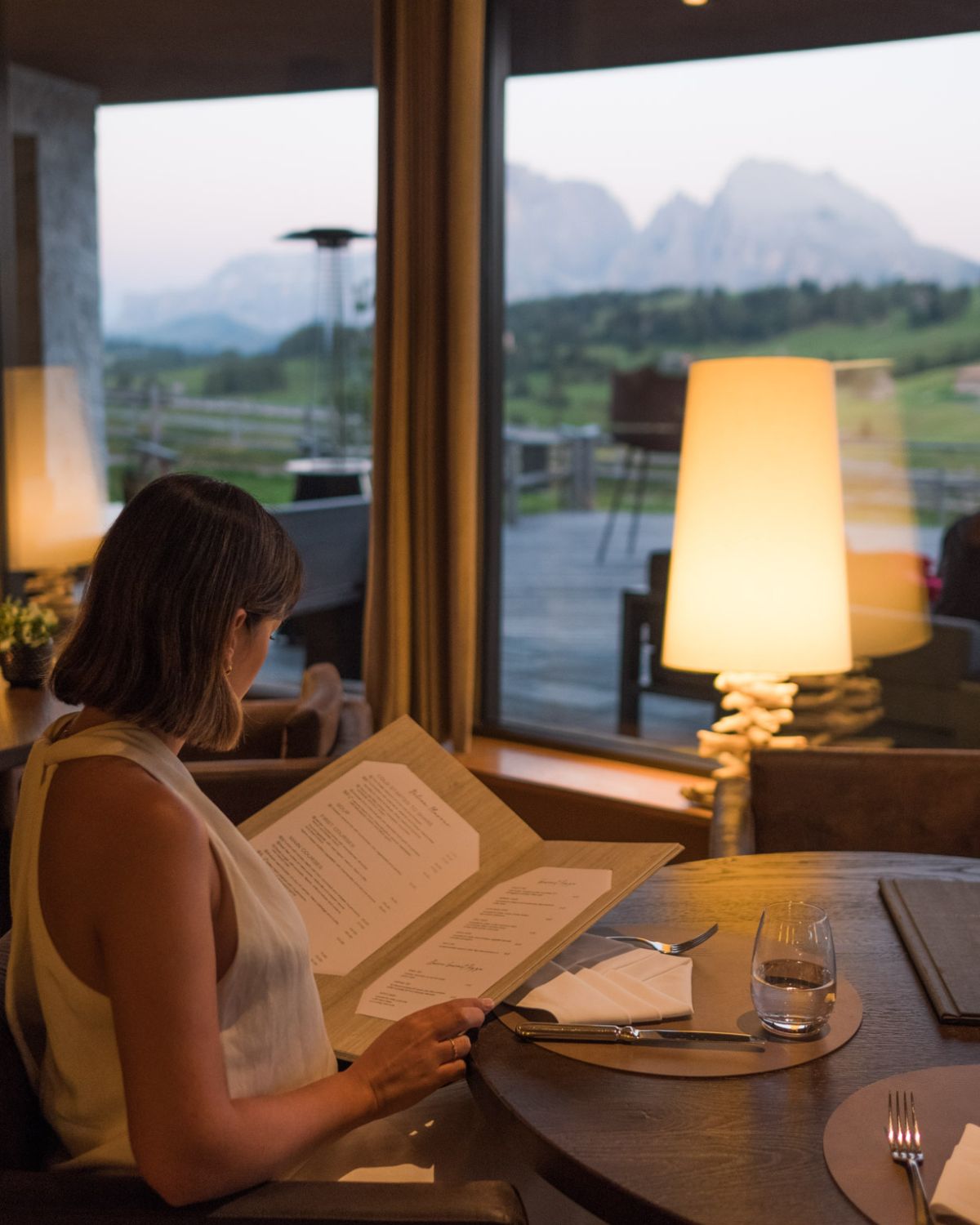 A lady in a white dress looking at a menu in a warm light restaurant setting, overlooking the mountains and a green hill.