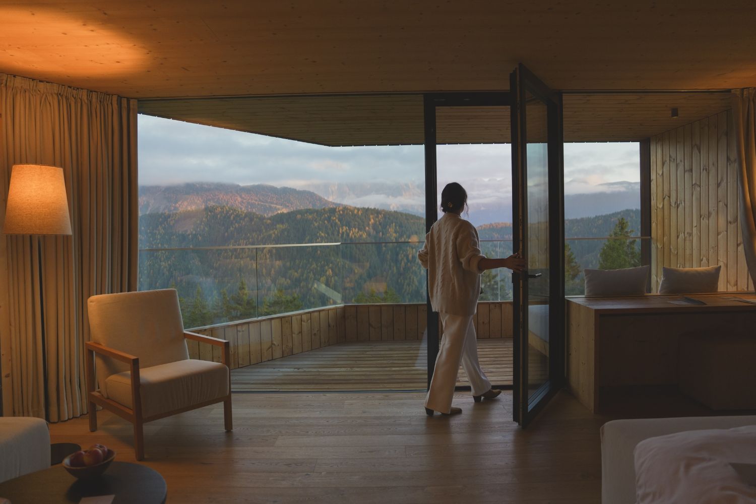 A lady walking inside a hotel room with a cabin style, wood ceilings, floors and walls, overlooking a wide view of the mountains in the back.