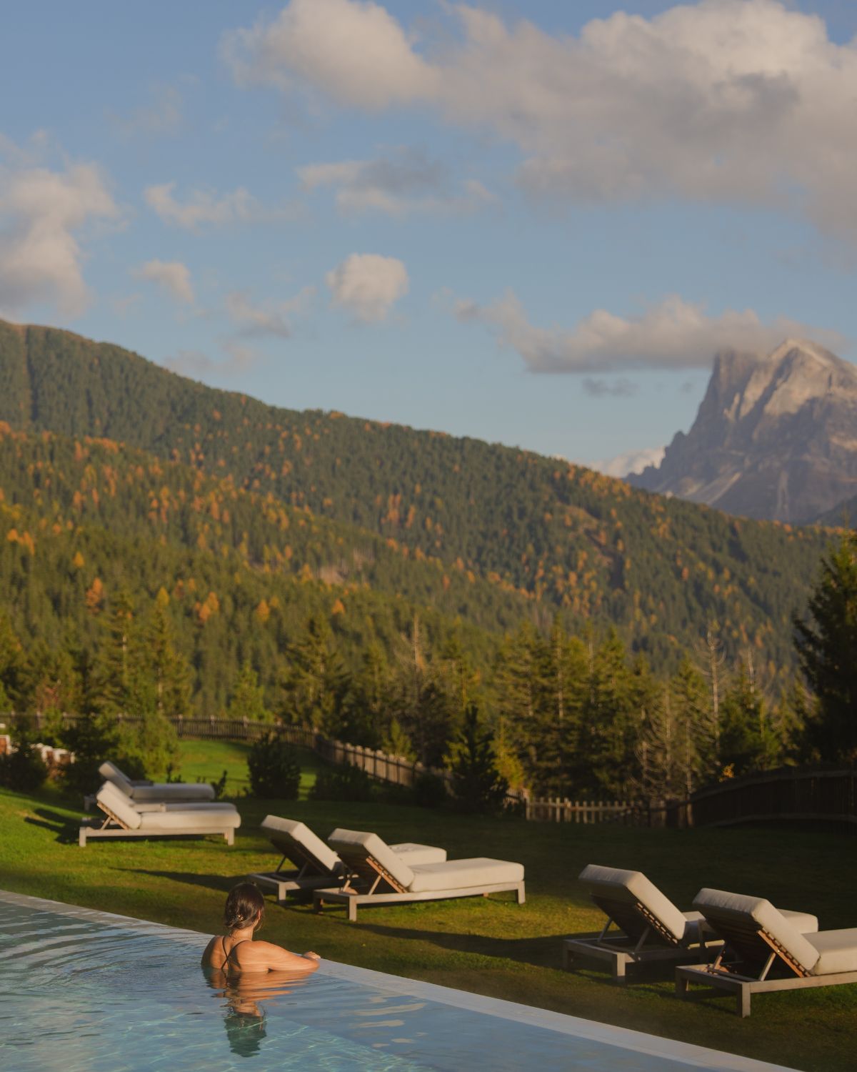 A lady inside an outdoor pool, overlooking green and snowy mountains, in front of sunbeds.