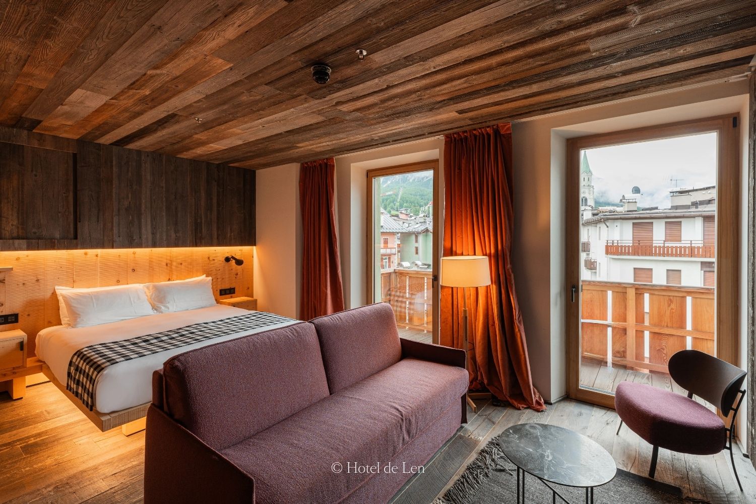 A hotel room, with wooden floors, walls and ceilings, a white linen bed, a mauve sofa and chair, next to tall doors facing buildings and a mountain.