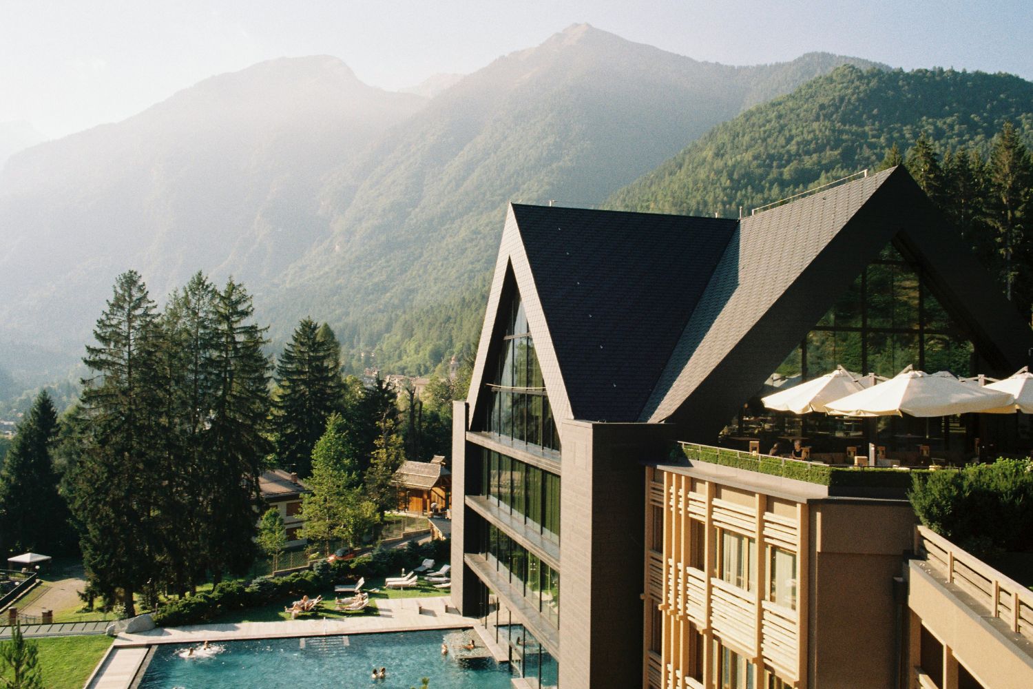 A cabin like hotel façade, with large windows, an outdoor pool and lounge space with white sunshades, surrounded by high green mountains.