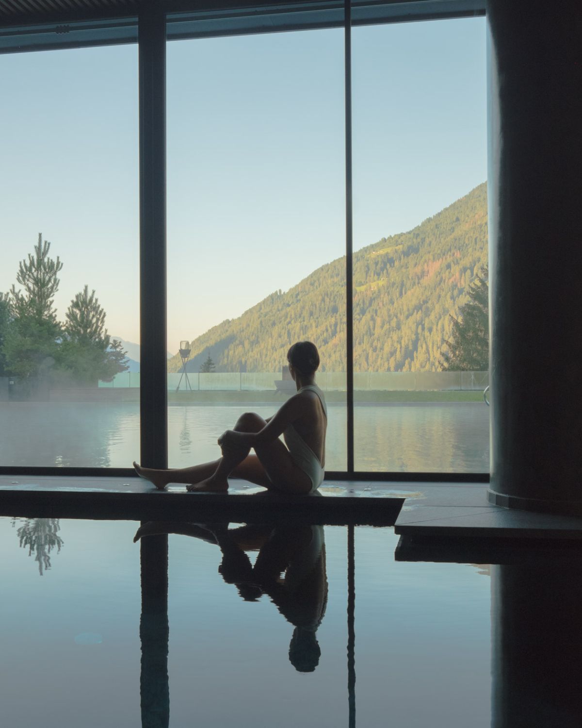 Lady in a white swimsuit, next to an indoor pool, overlooking a green mountain in front of large windows.