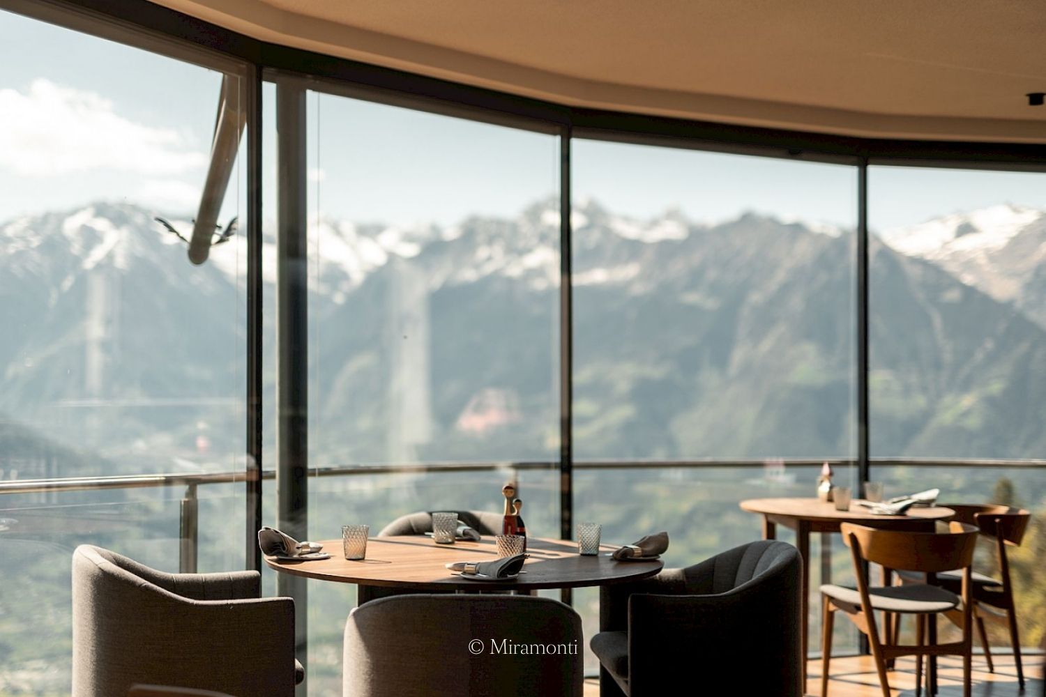 A restaurant setting with dark grey and wood furniture, next to wide windows, overlooking a snowy mountain.