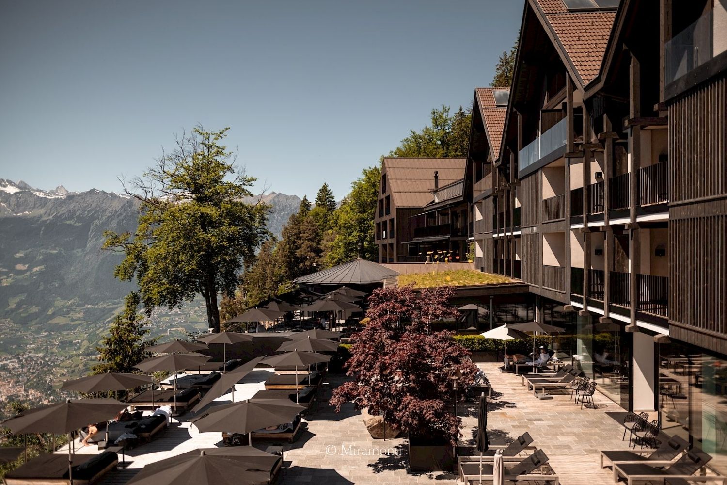 A cabin like hotel façade, with large windows, and an outdoor lounge space with black sunshades, surrounded by high mountains.