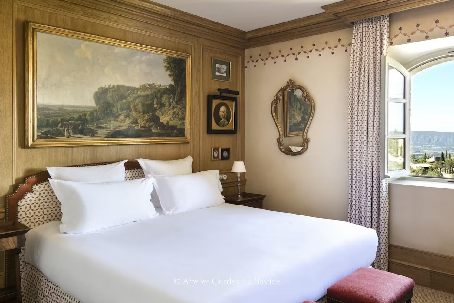 Wood paneling wall with a oil landscape painting within a gold frame, above a white linen kind bed, next to an antique gold mirror and a window facing a green landscape, with trees and bushes.