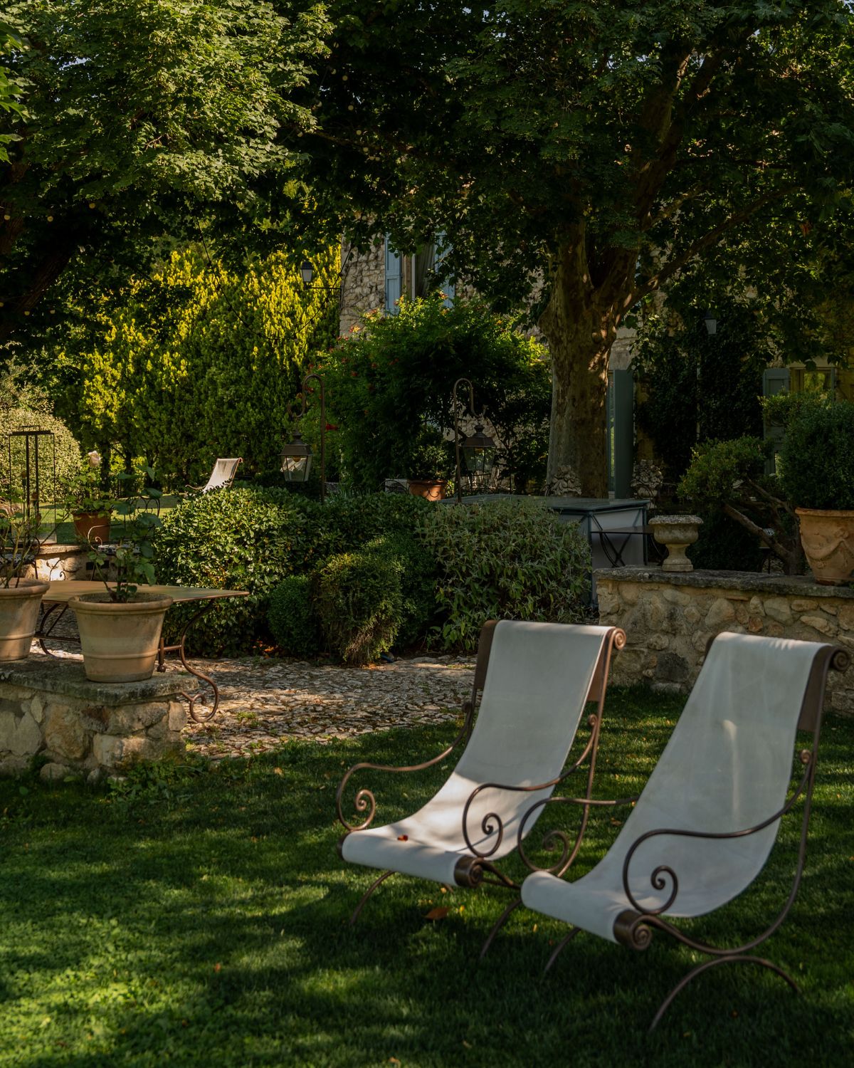 Dark iron and white linen outdoor chairs, on grass, in front of bushes, a high tree, a low stone wall and a light blue shutters house.