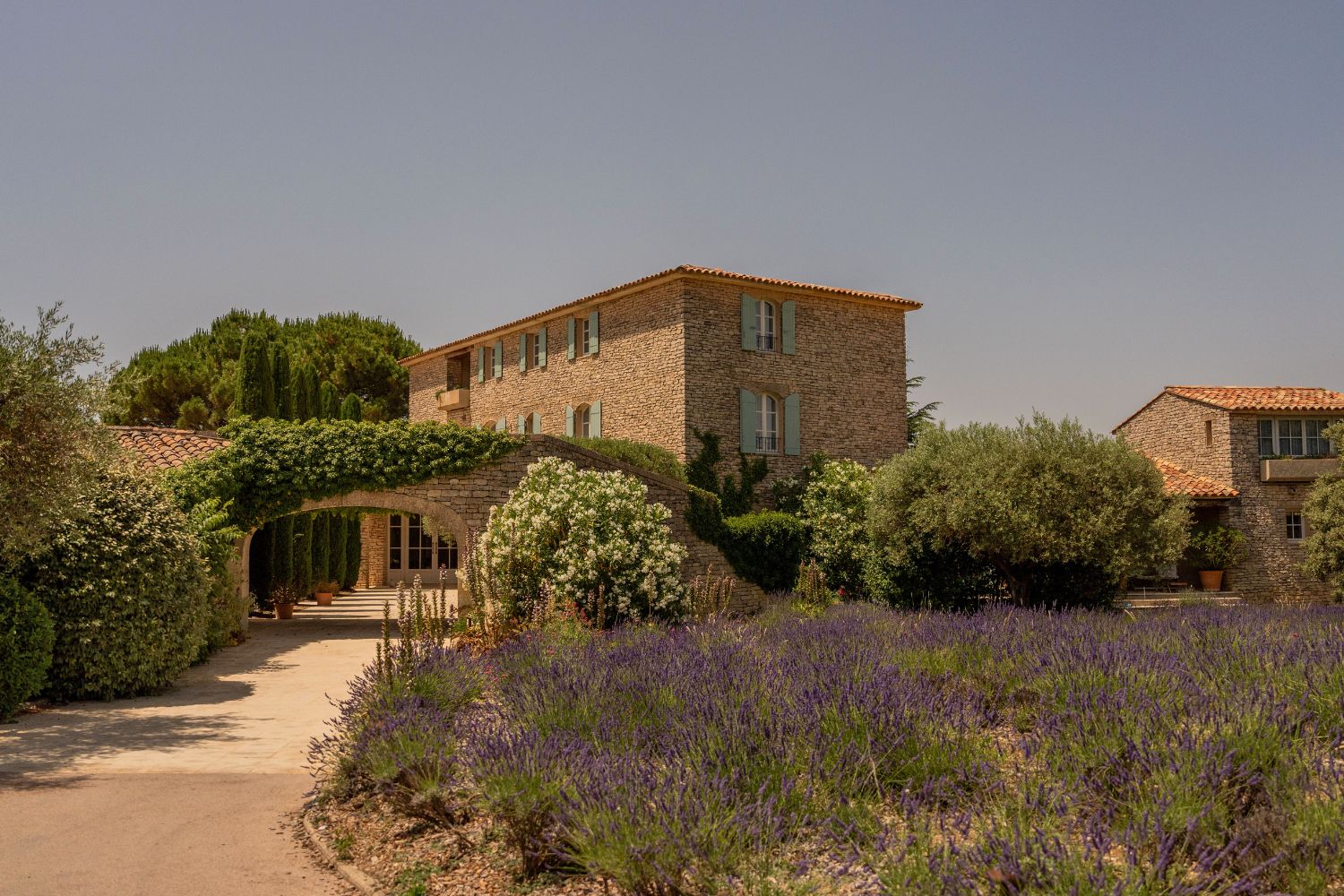 Cobblestone house, with light blue shutters, an archway with climbing trees, an abundant garden in front with lavender bushes.