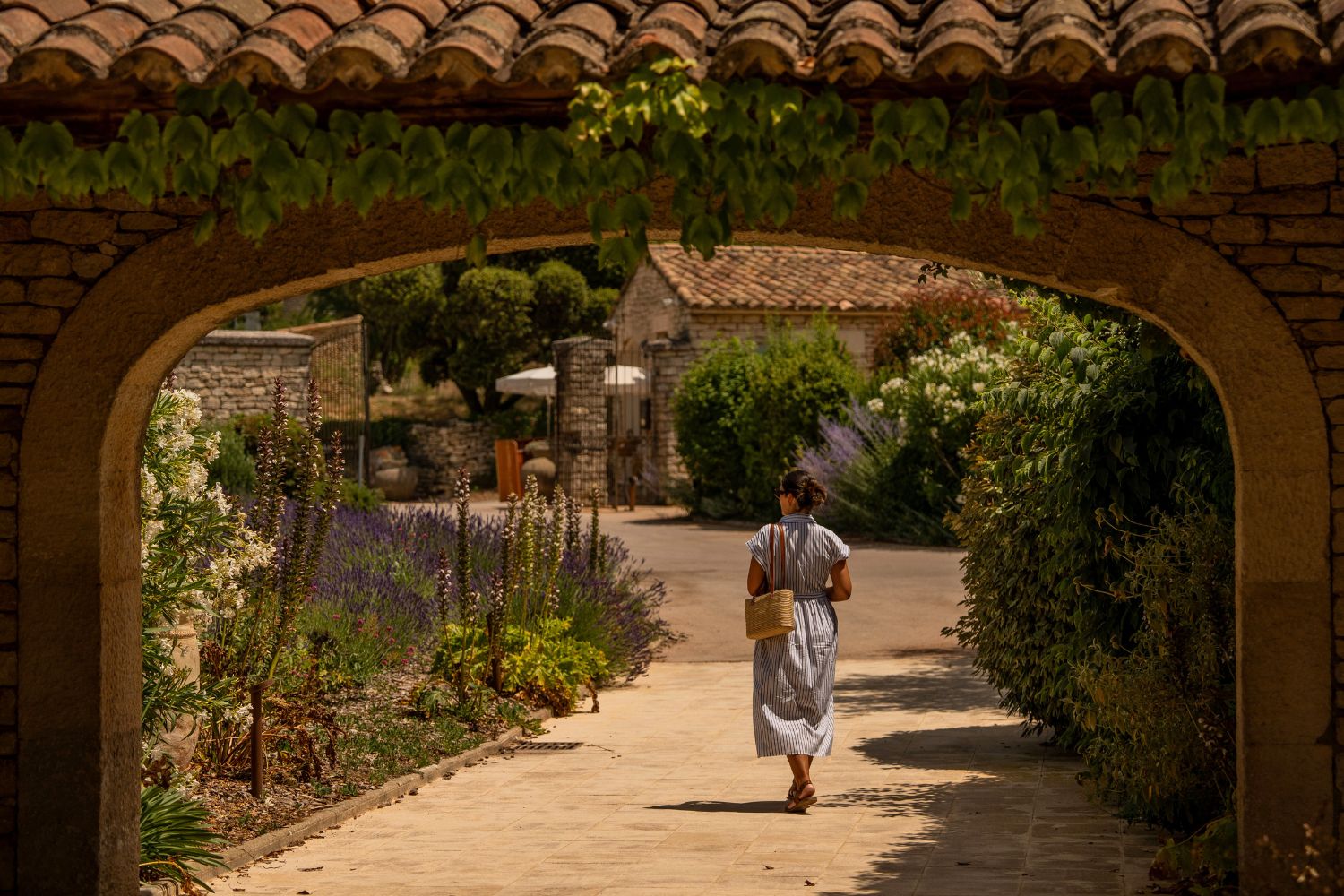 Woman with a stripe white and blue dress, walking on a dirt road, next to an abundant garden, with a cobblestone house in the background.