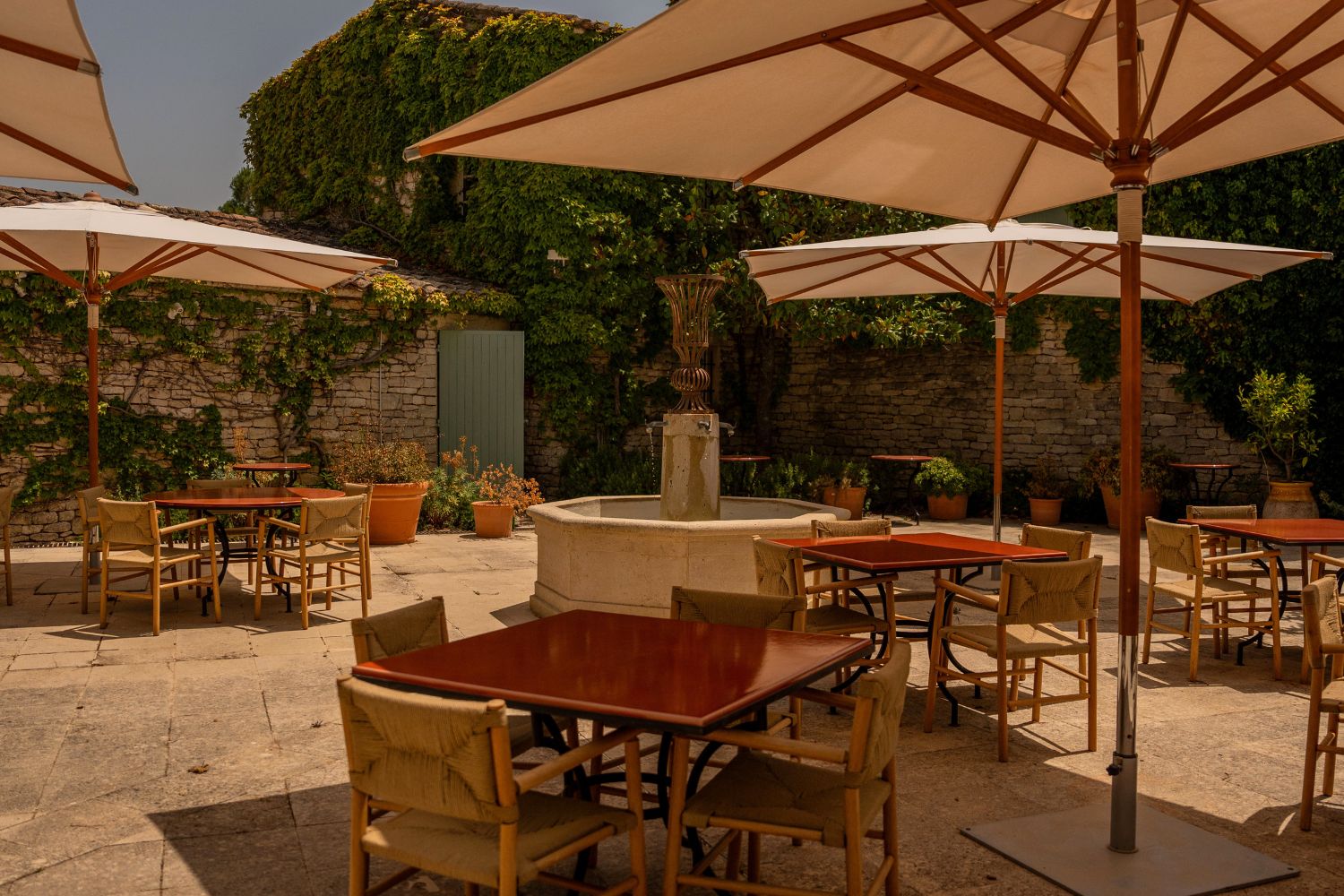 Outdoor restaurant setting, with red square tables, light wood chairs and sunshades, a fountain and climbing plants on a stone wall.