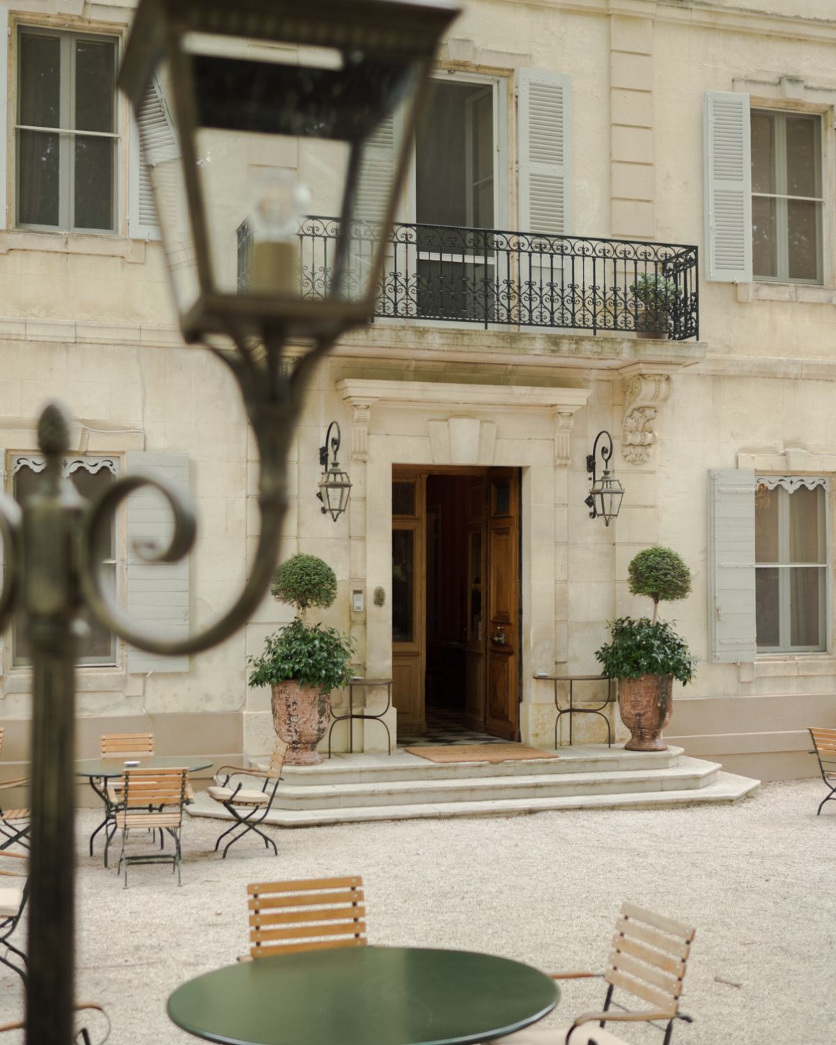 Cream façade, with light blue shutters, a wooden entrance door, iron and wooden outdoor furniture and black iron lamps.