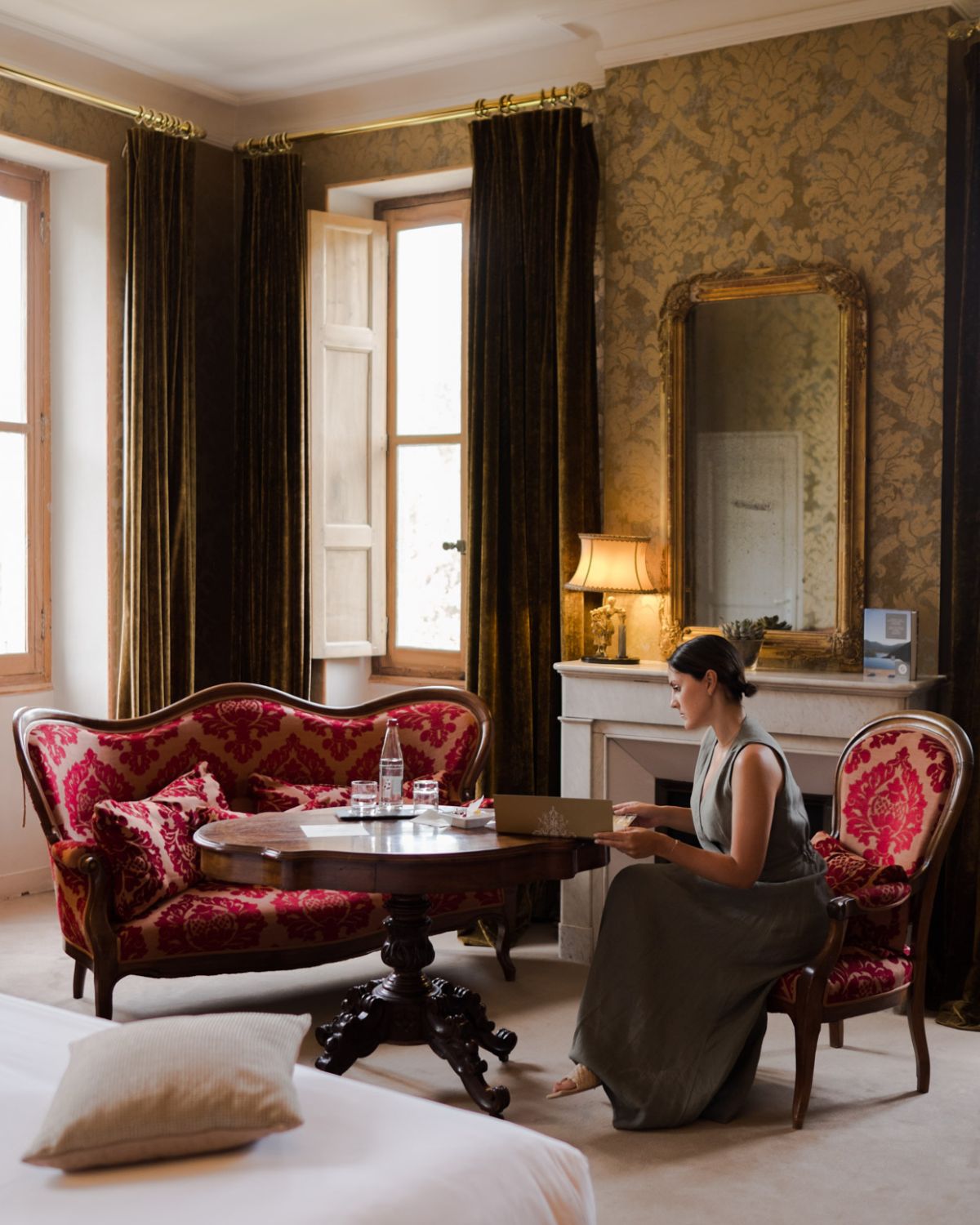 Golden and red antique room. Golden wallpaper, dark green velvet curtains, a white fireplace with a gold antique mirror, behind a sage green dressed lady, standing on an antique red chair, next to a sofa and a wooden table.