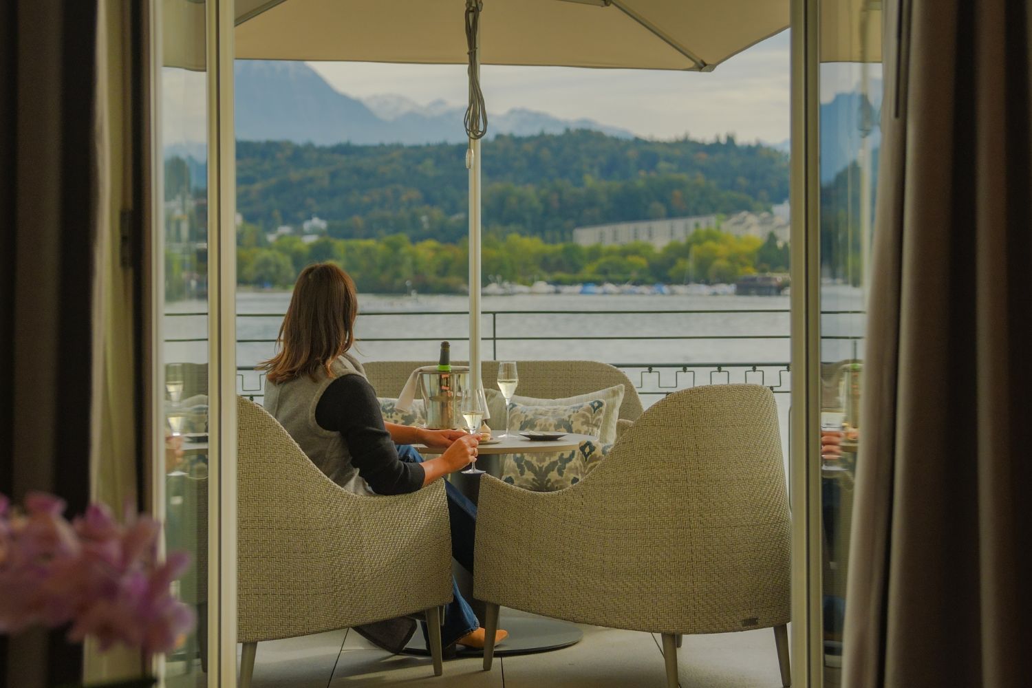 A lady sitting on an outdoor beige armchair, bellow a sunshade, overlooking a blue lake and mountains. The lady holds a glass of champagne.