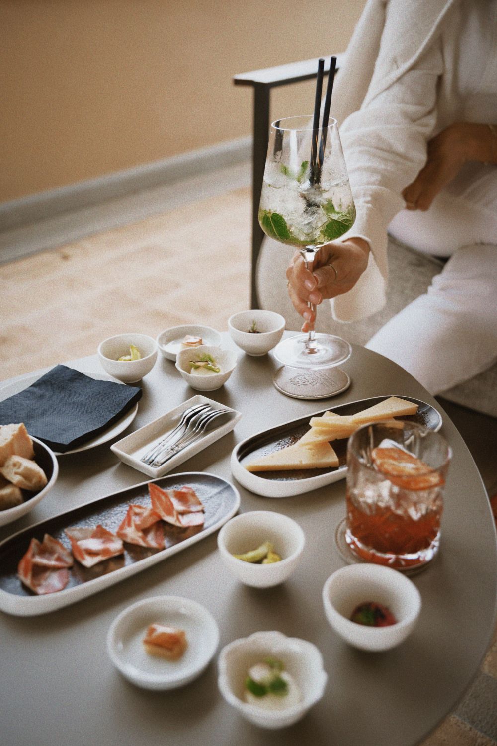 A centre dark table with appetizers on it and a white dressed lady on the corner with a glass on her hand.