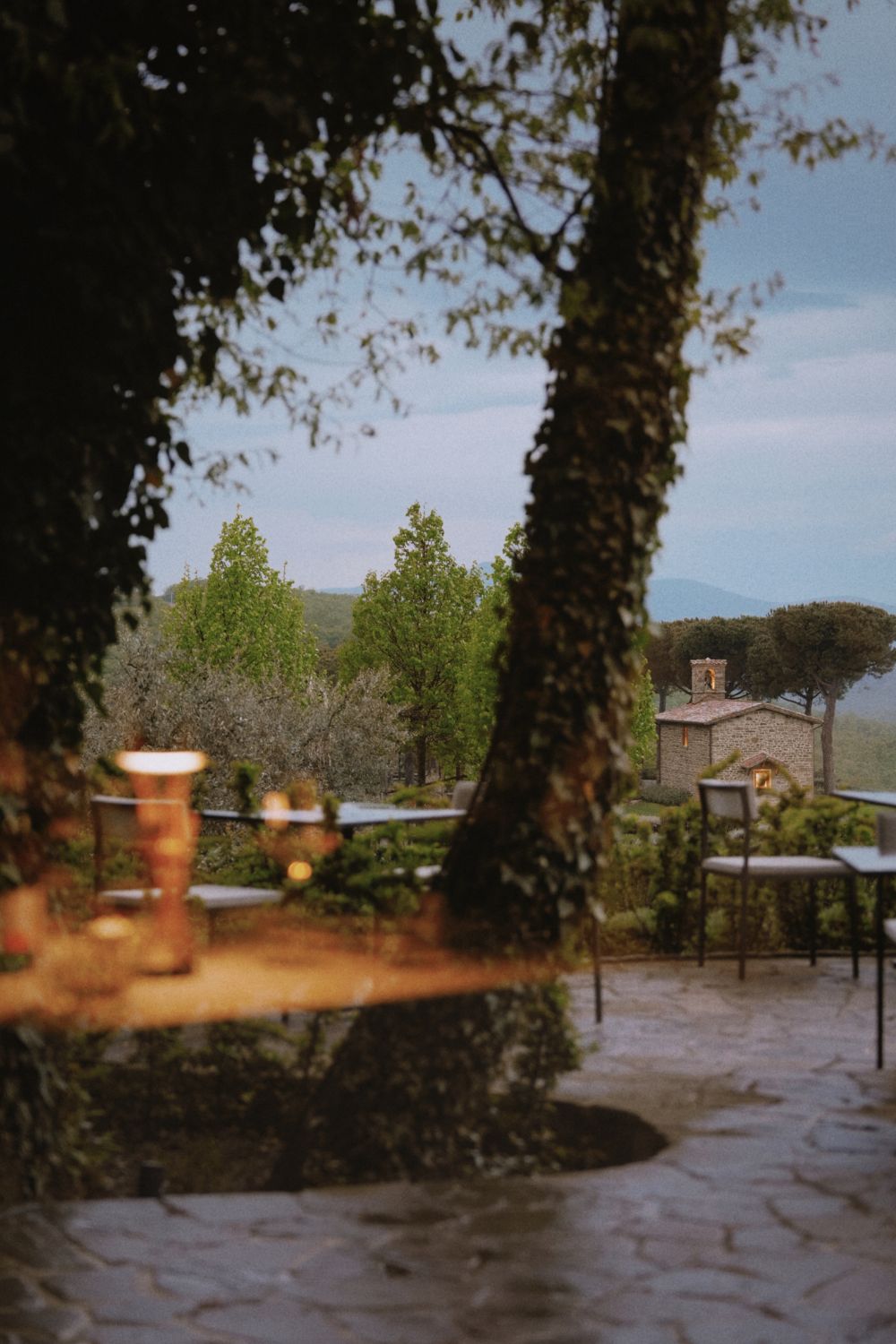 Iron outdoor set on top of a stone path in the evening with lights on, surrounded by a green scenery of trees, bushes, grass and a second cobbled stone house in the background.