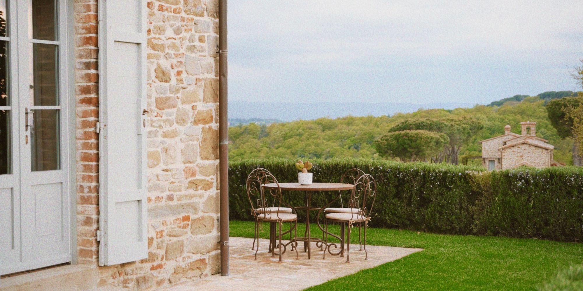Iron outdoor set on the corner of a cobbled stone house, with light grey shutters, surrounded by a green scenery of trees, bushes, grass and a second cobbled stone house in the background.