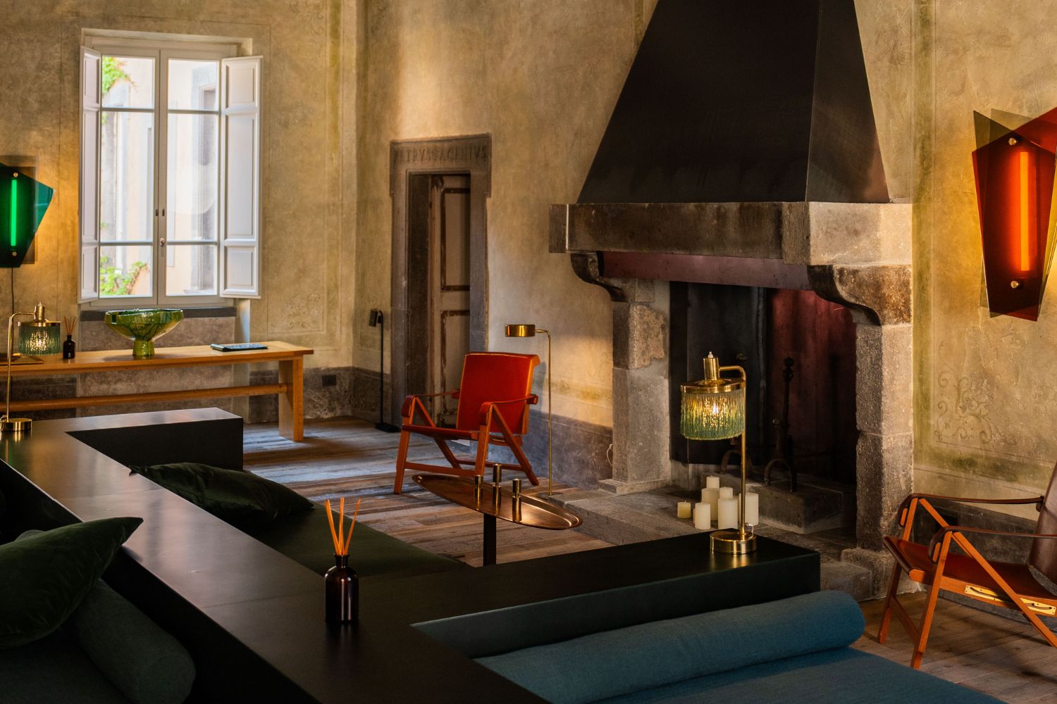 Traditional fireplace, with dark contemporary sofa in front, two red leather armchairs, a green and red lamps on the lime wash old walls.