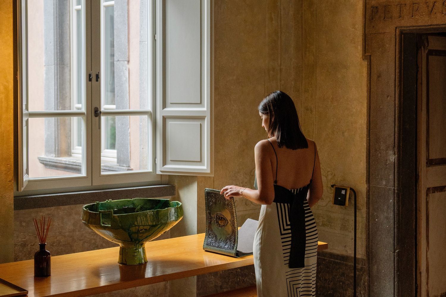 A lady with a geometric patterned dress next to a light wooden table, in front of a shuttered window, inside an old living room with lime wash walls.