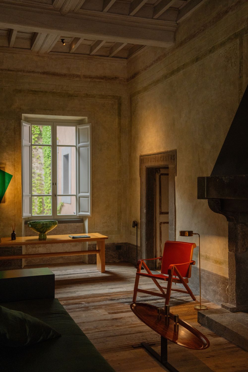 Traditional fireplace, with dark contemporary sofa in front, one red leather armchairs, a green lamp on the lime wash old wall and a wooden table.