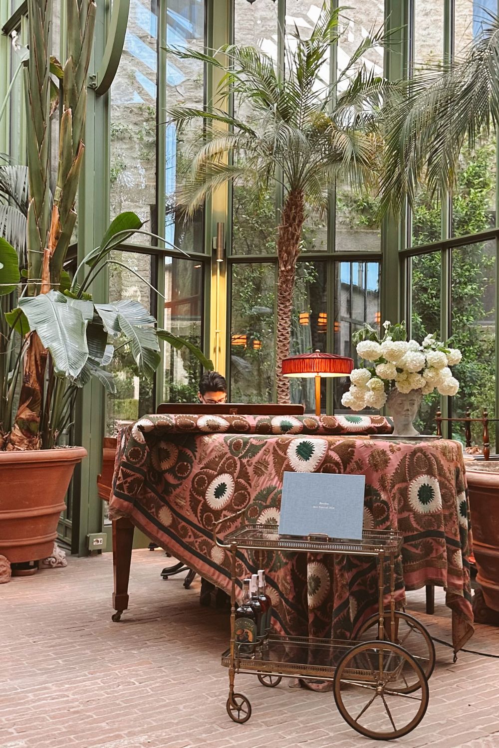 Wooden piano covered with a green and muted red blanket, inside a green glass house, with palm trees.