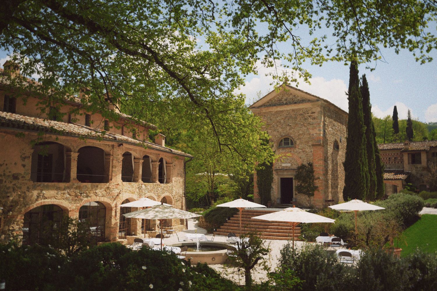 Cobbled stone buildings with archways, a fountain, white sunshades and green surroundings.
