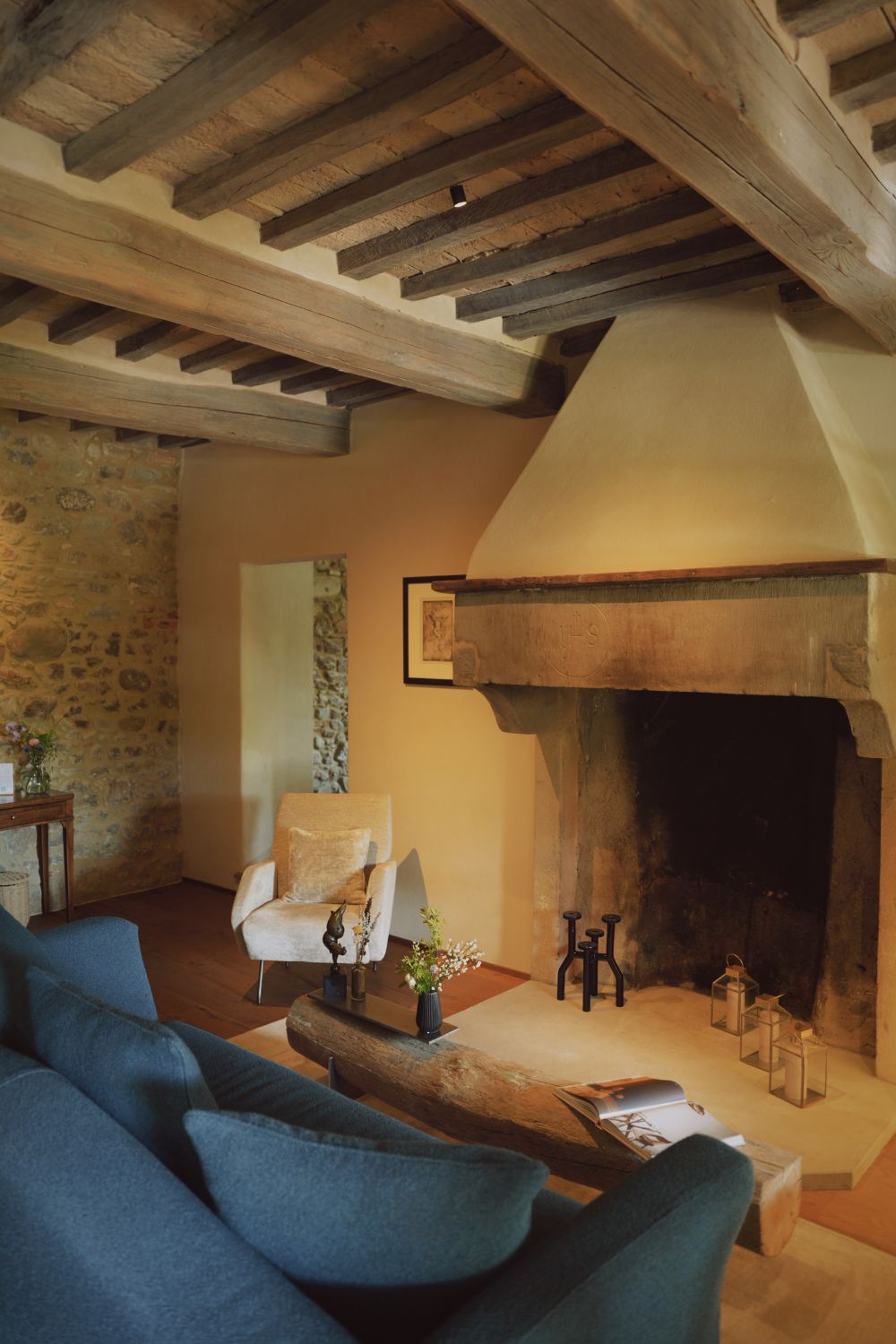Old fireplace, in front of a blue sofa, below a wooden beam ceiling.