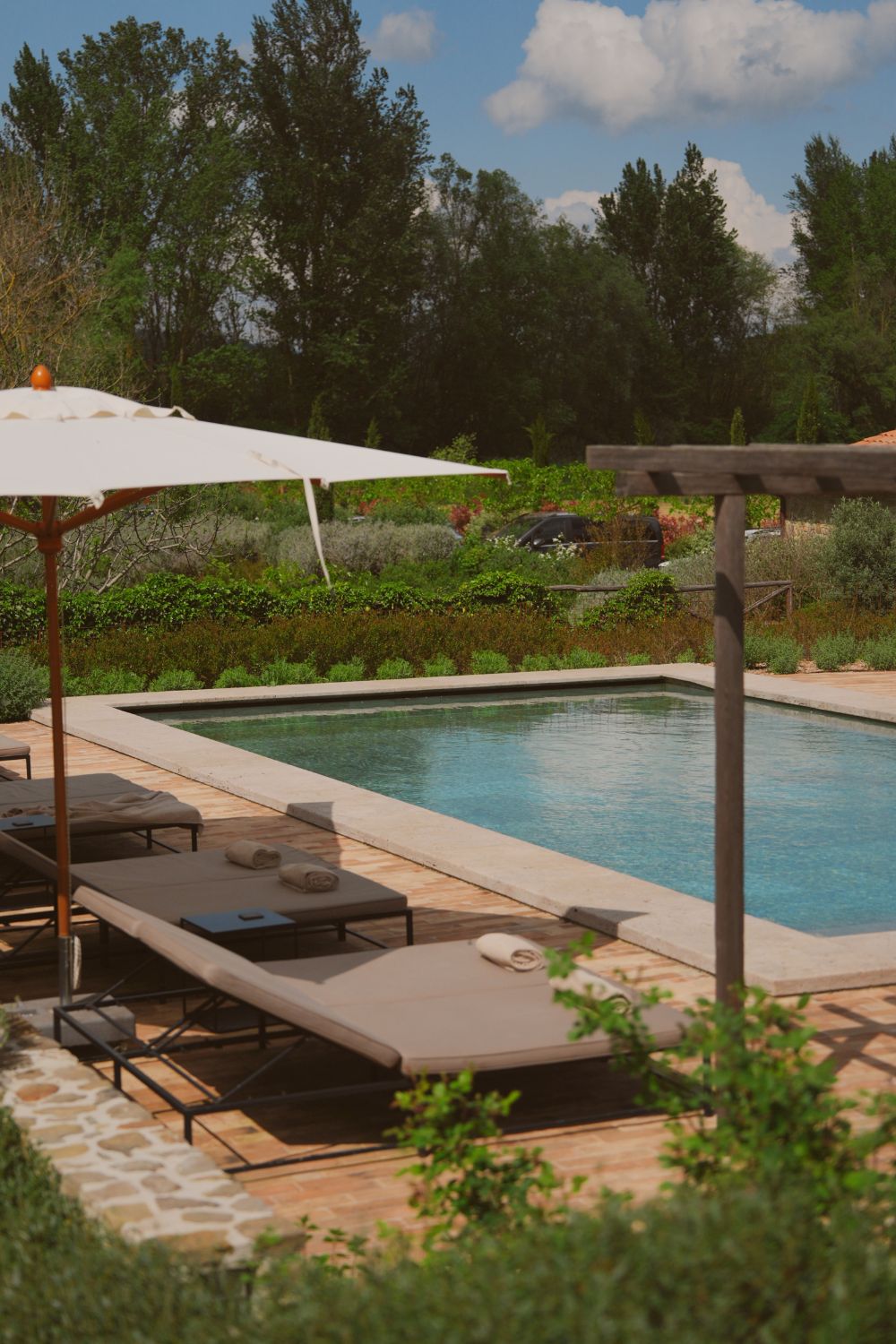 Blue pool, next to a white sunshade and grey sun beds, surrounded by green trees and bushes.