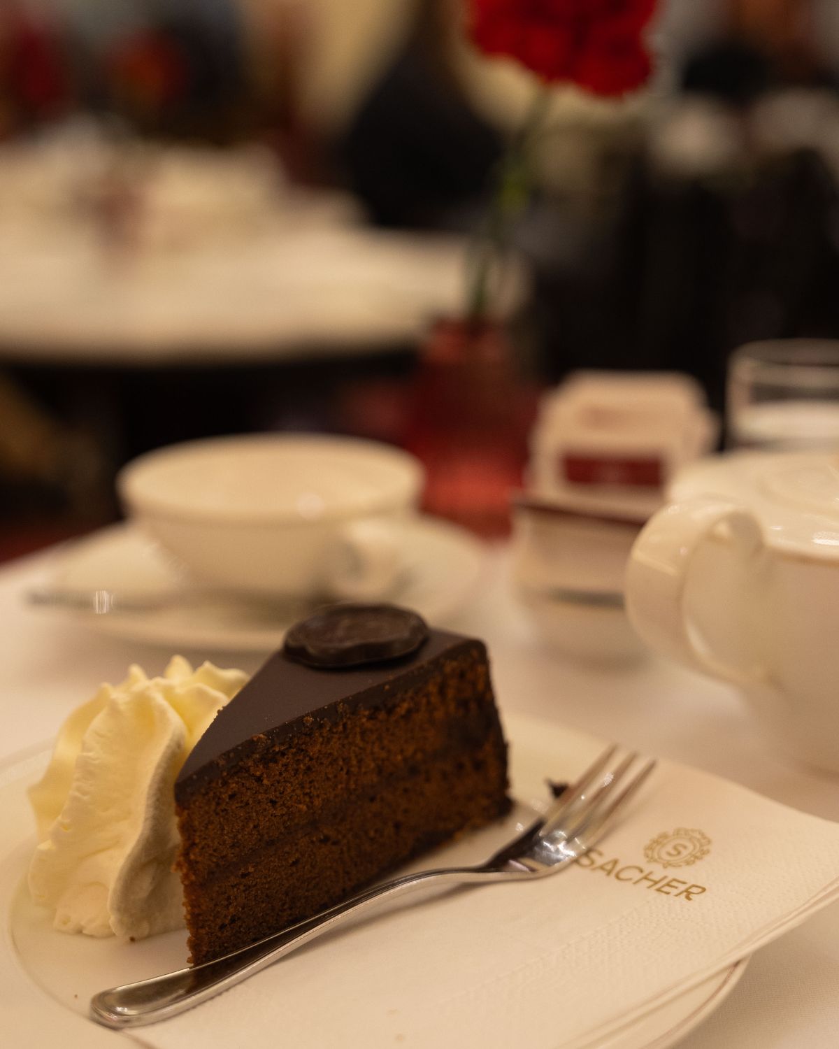 A dark chocolate cake slice with cream on the side on a white tablecloth.
