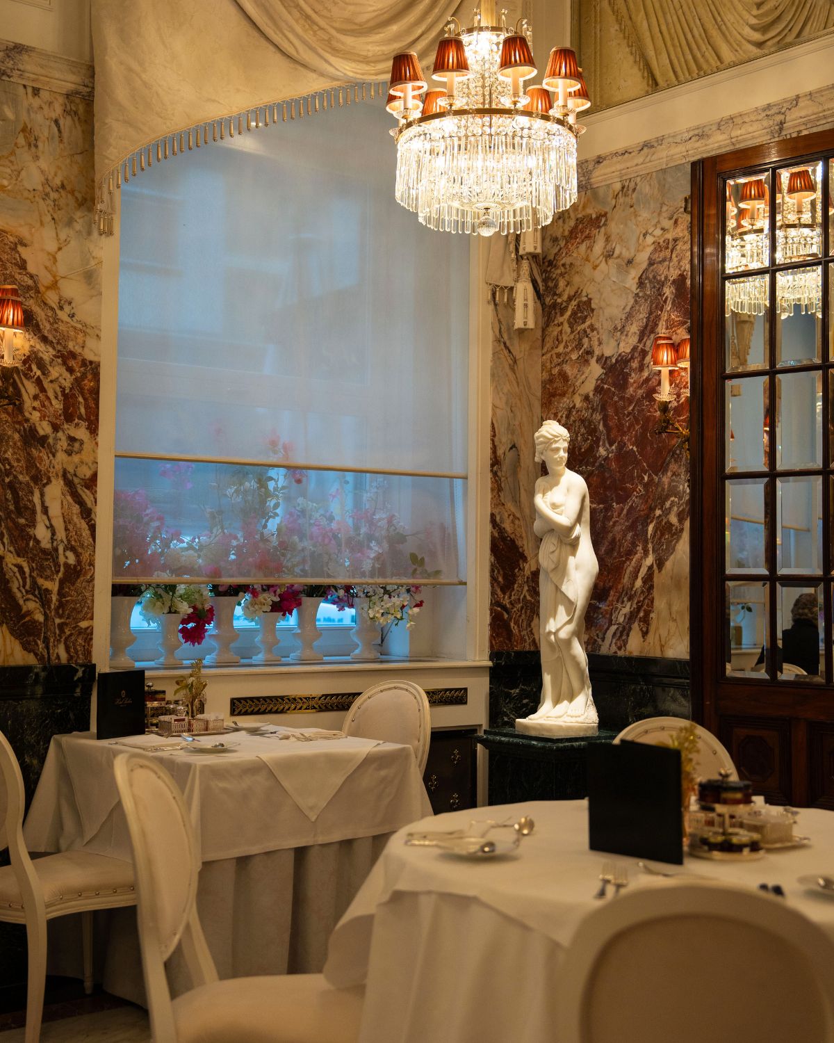 A classical marble dining room, with a crystal chandelier, a white sculpture, chairs and tablecloths.