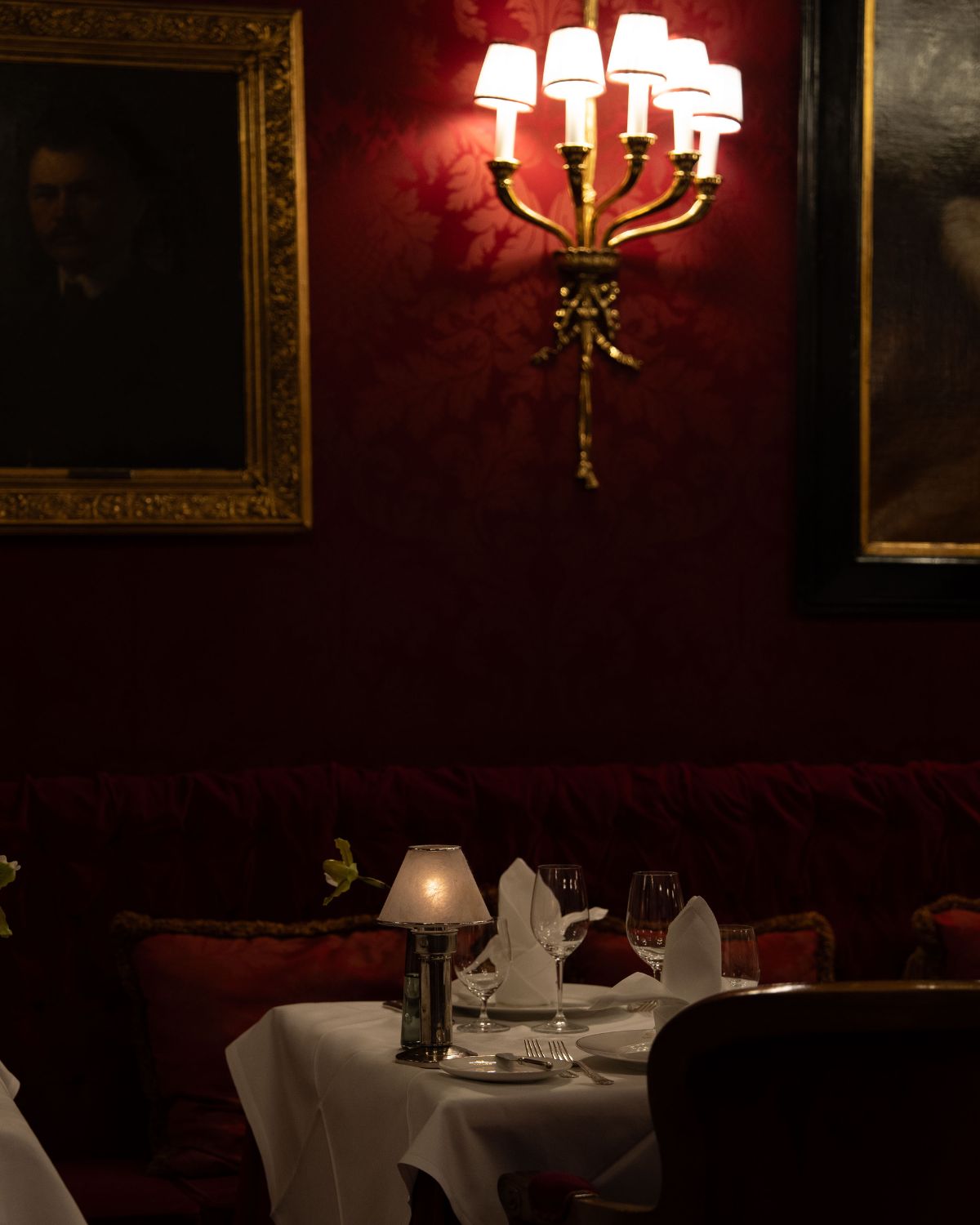 A classical dark red and golden dining room setting, with golden frames, wall lamps and white tablecloths.