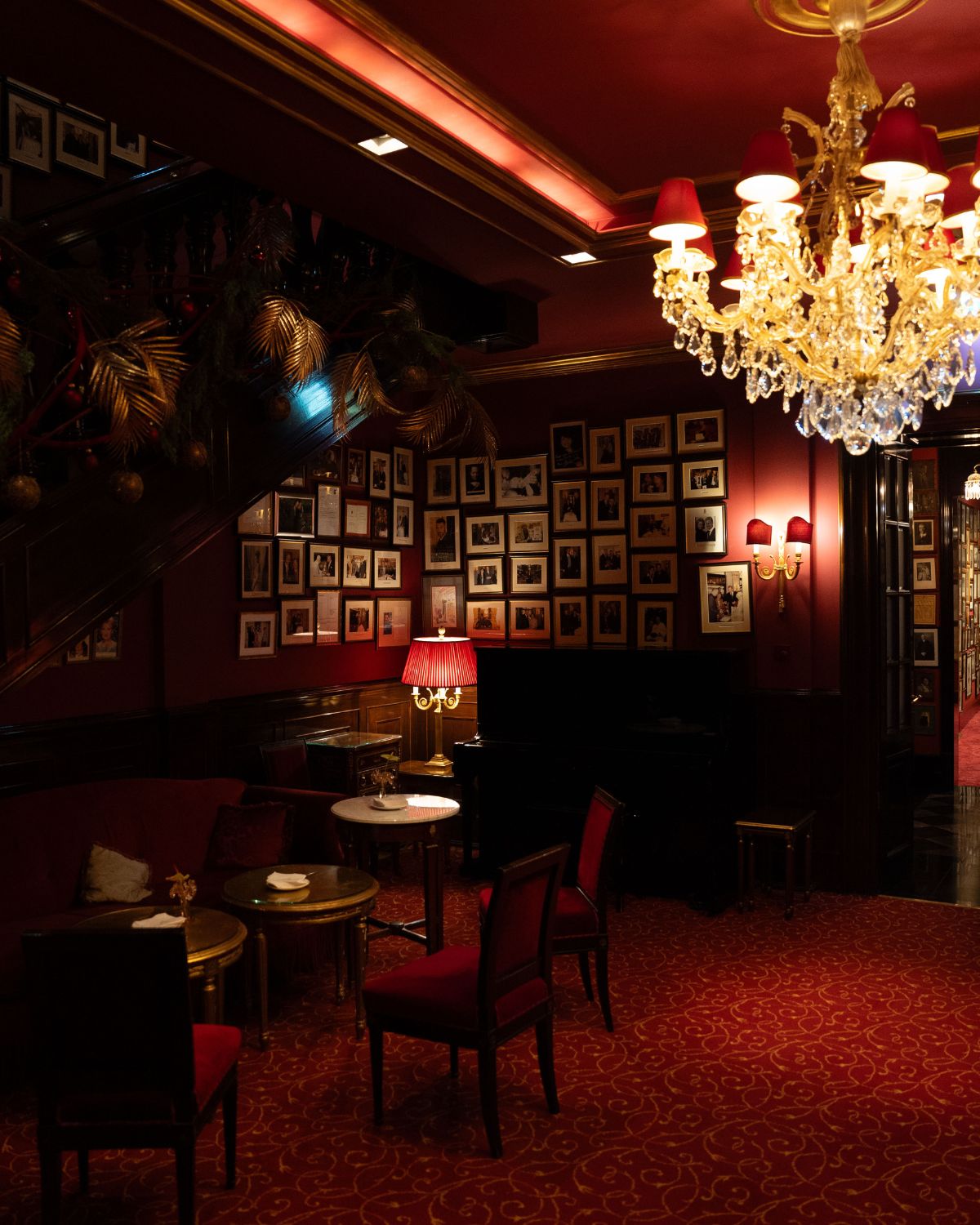 A classical dark red lounge space, with red furniture, a crystal chandelier, multiple frames on the wall and a Christmas adorned staircase.