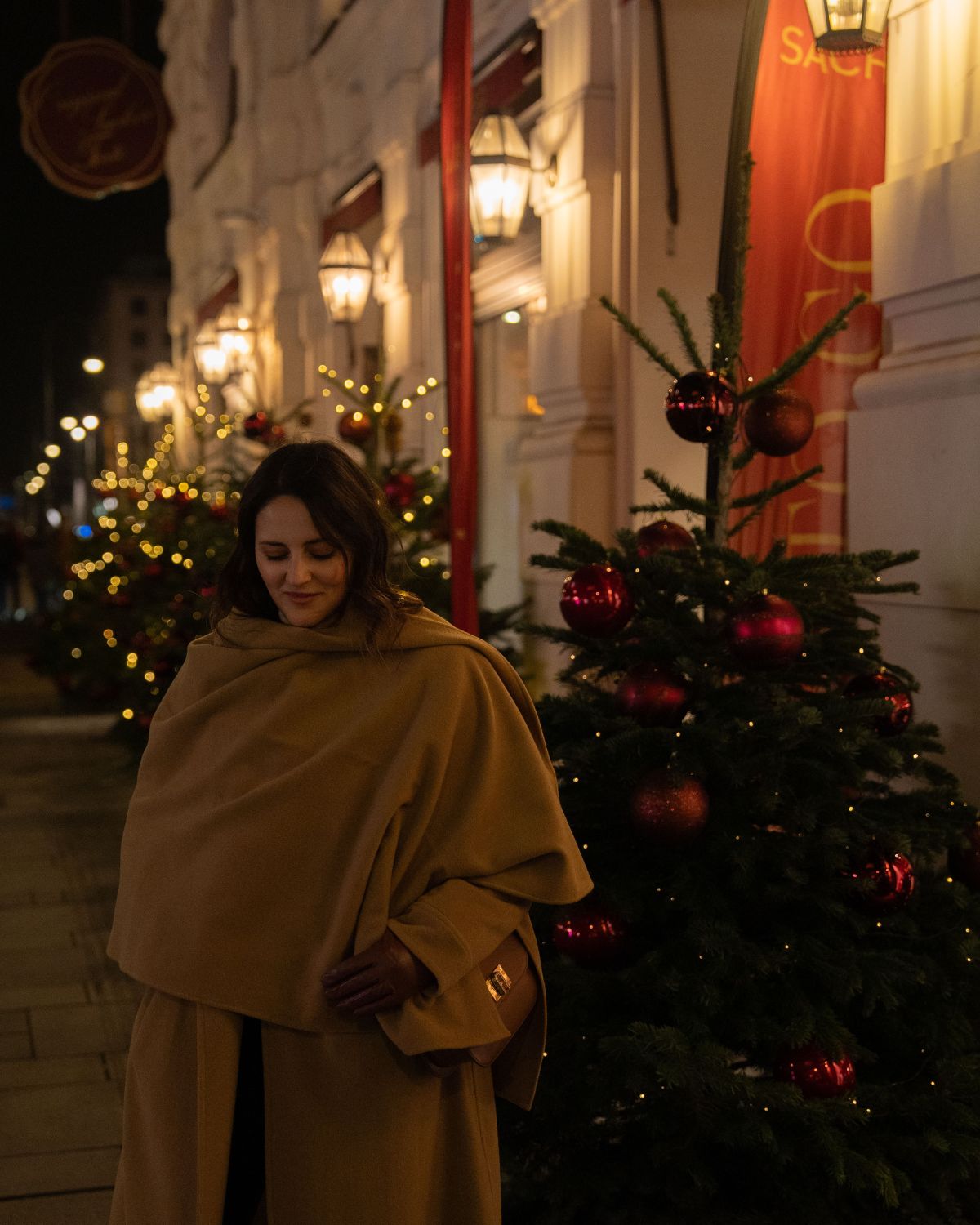 A lady with a cream coat and scarf in the street at nighttime, next to a Christmas tree and a classical building façade.